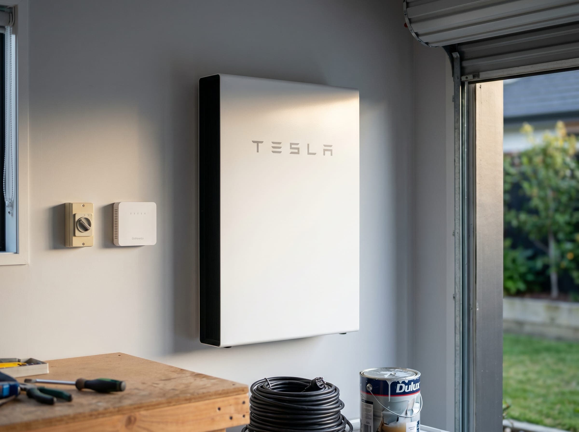 A Tesla Powerwall 3 mounted on the wall of a New Zealand homeowner's garage, next to a small electrical isolator