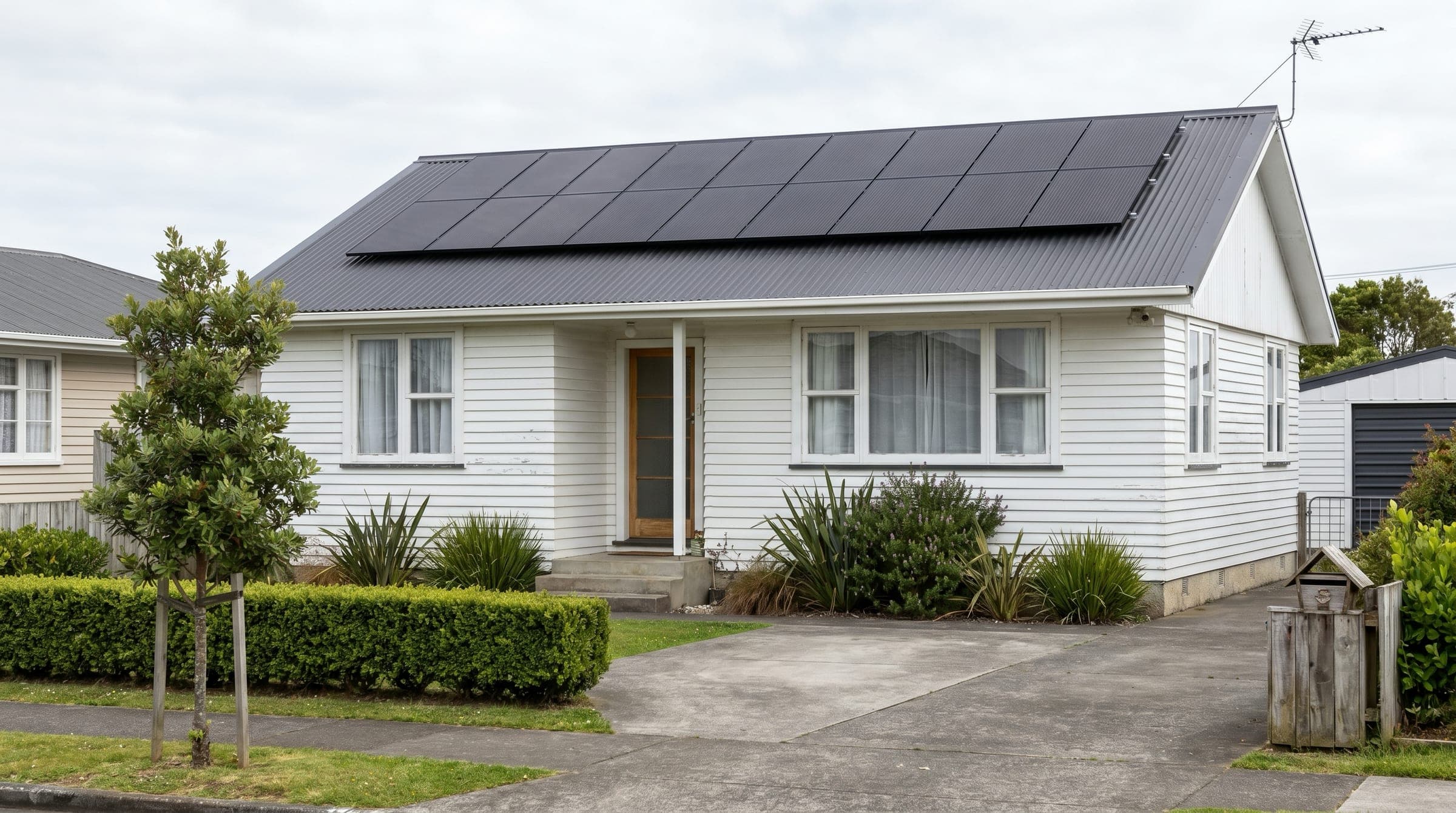 Documentary photo of a modest South Auckland white-weatherboard suburban home with a tidy row of all-black solar panels on its charcoal corrugated steel roof