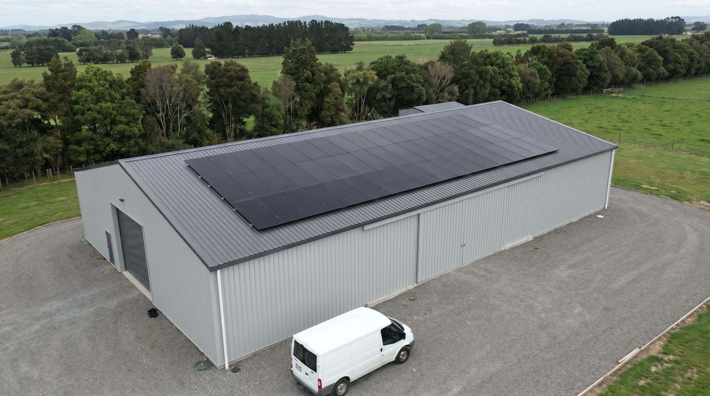 Documentary photo of a rural NZ commercial shed with a large all-black solar array on its charcoal corrugated steel roof, a white tradie van parked in front, shelterbelt trees and rolling pasture in the background