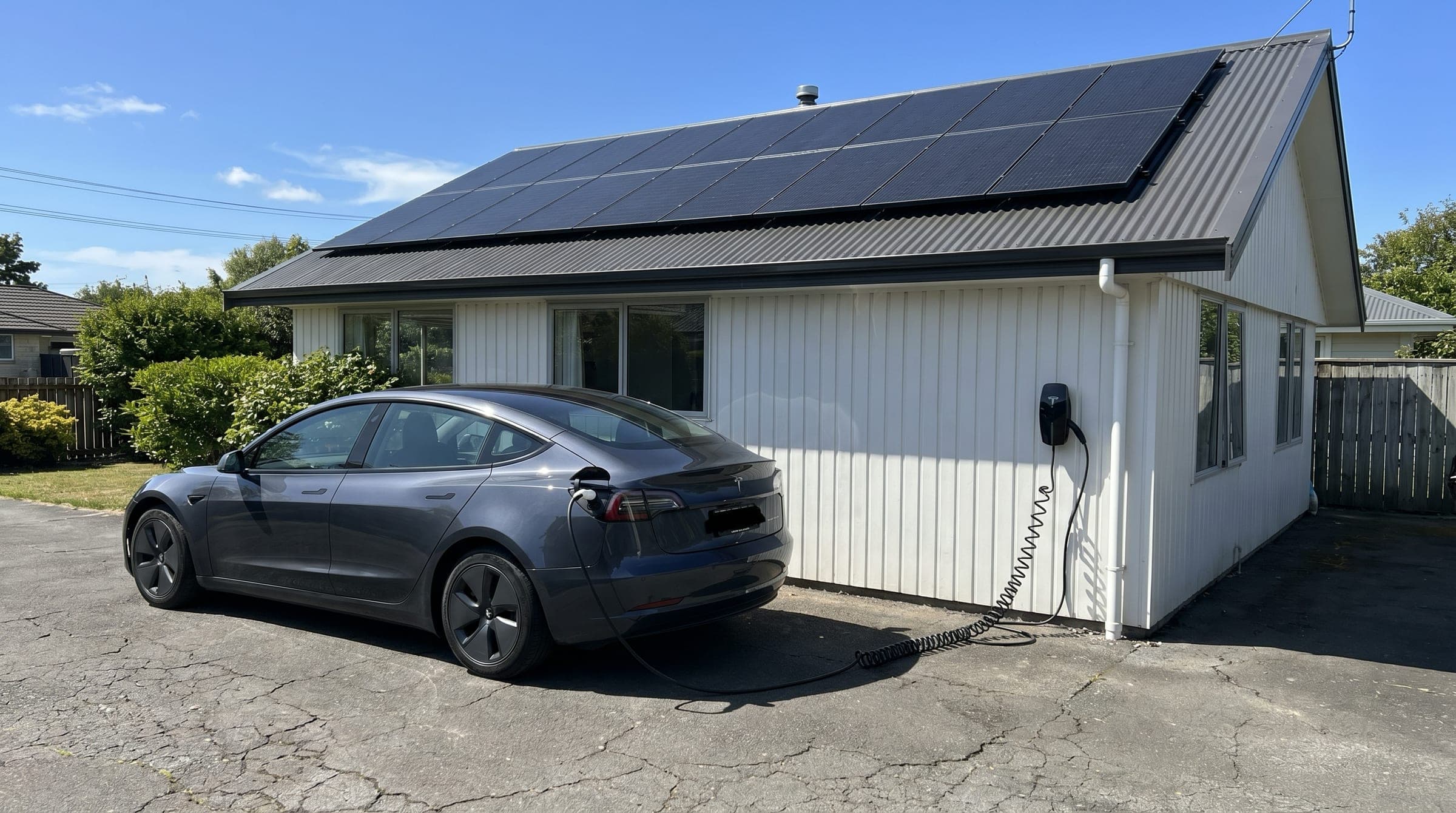 A modern electric vehicle plugged into a wall-mounted home charger on a Kiwi driveway, with all-black solar panels visible on the home's roof above