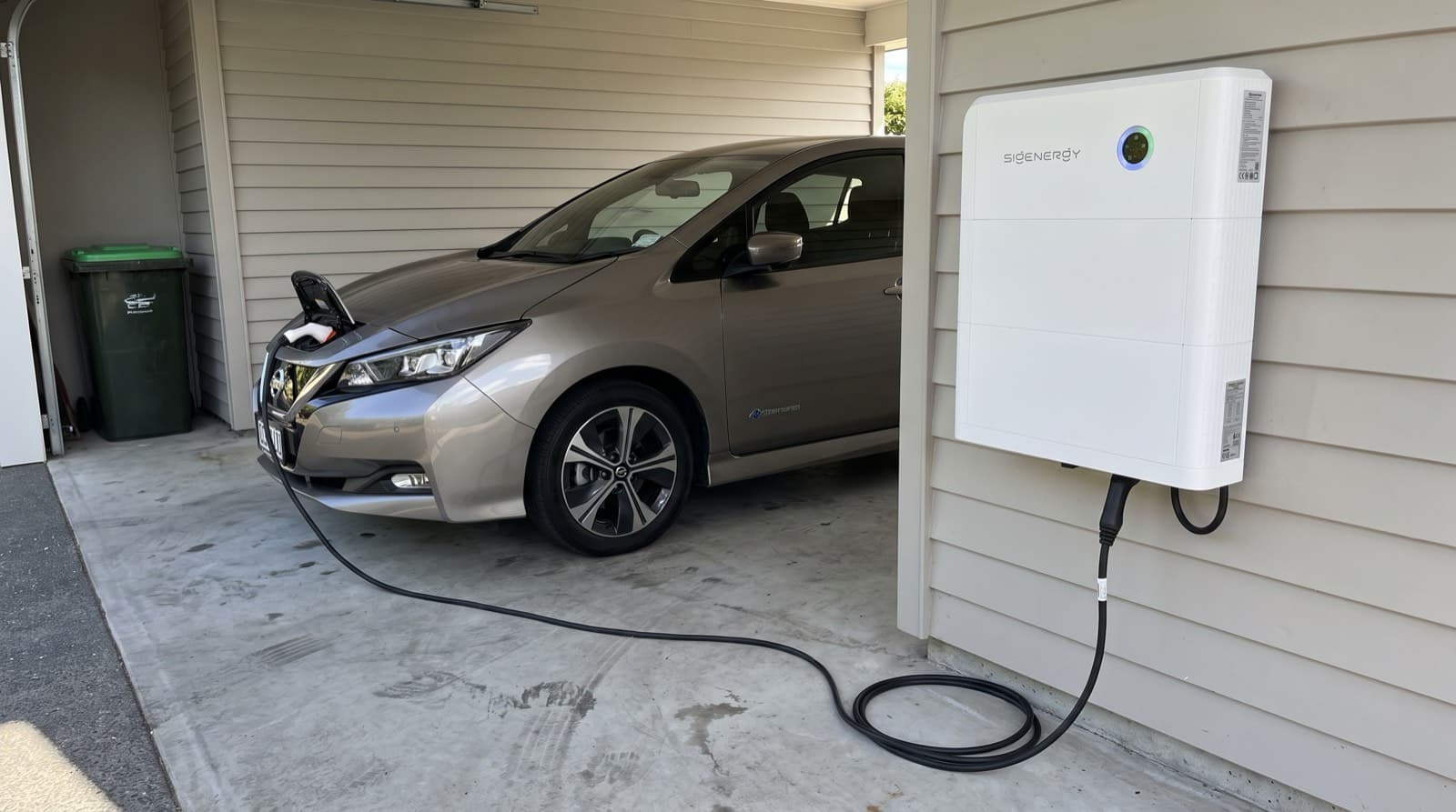 A white Sigenergy SigenStor battery stack mounted in an ordinary New Zealand carport with a charging cable running to an EV hatchback