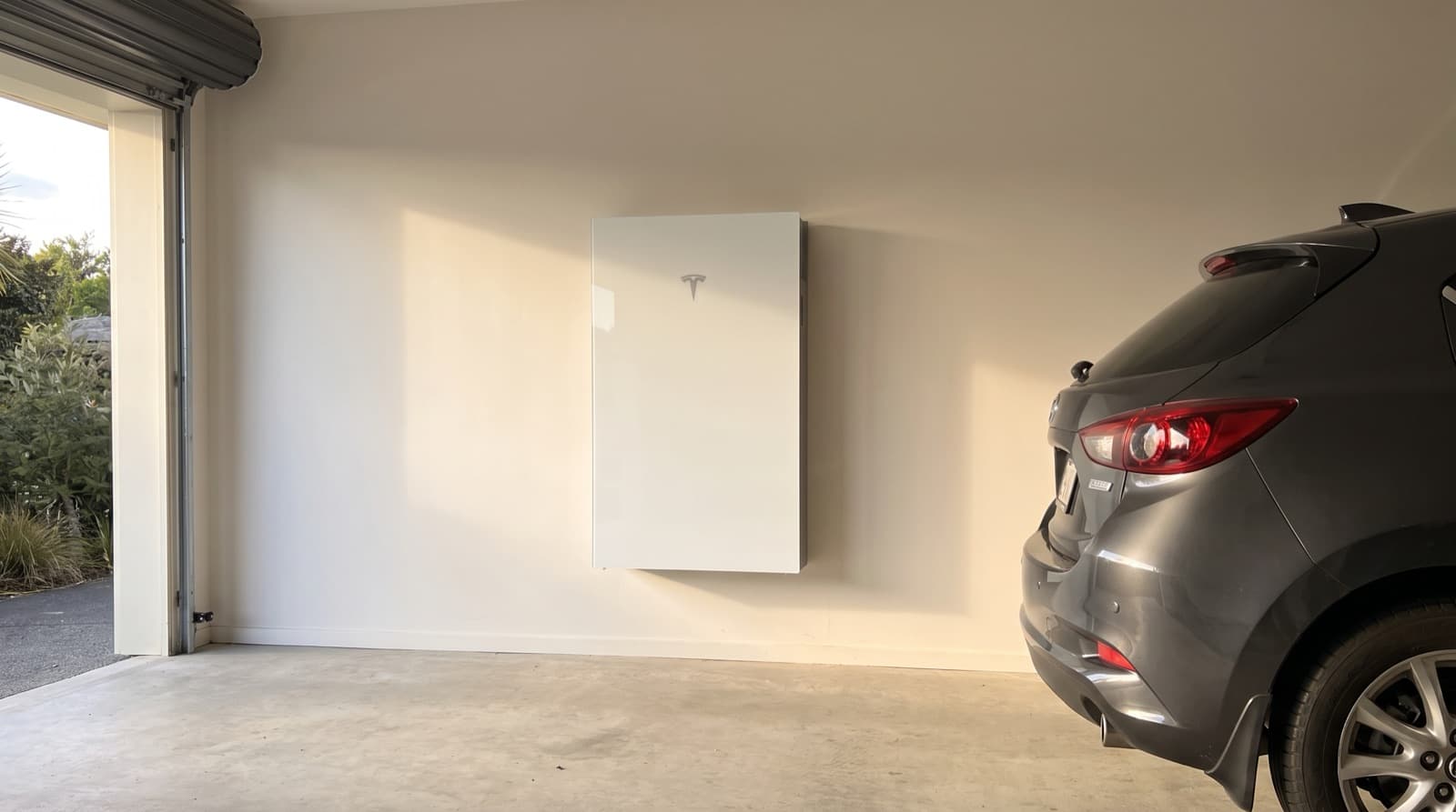A white Tesla Powerwall 3 mounted on the garage wall of a New Zealand home in evening light, with the family car parked beside it