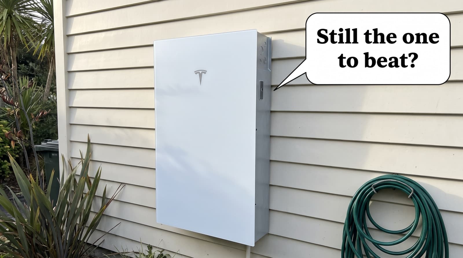 A white Tesla Powerwall 3 mounted on the cream weatherboard wall of a New Zealand home beside a coiled garden hose, speech bubble asking if it is still the one to beat