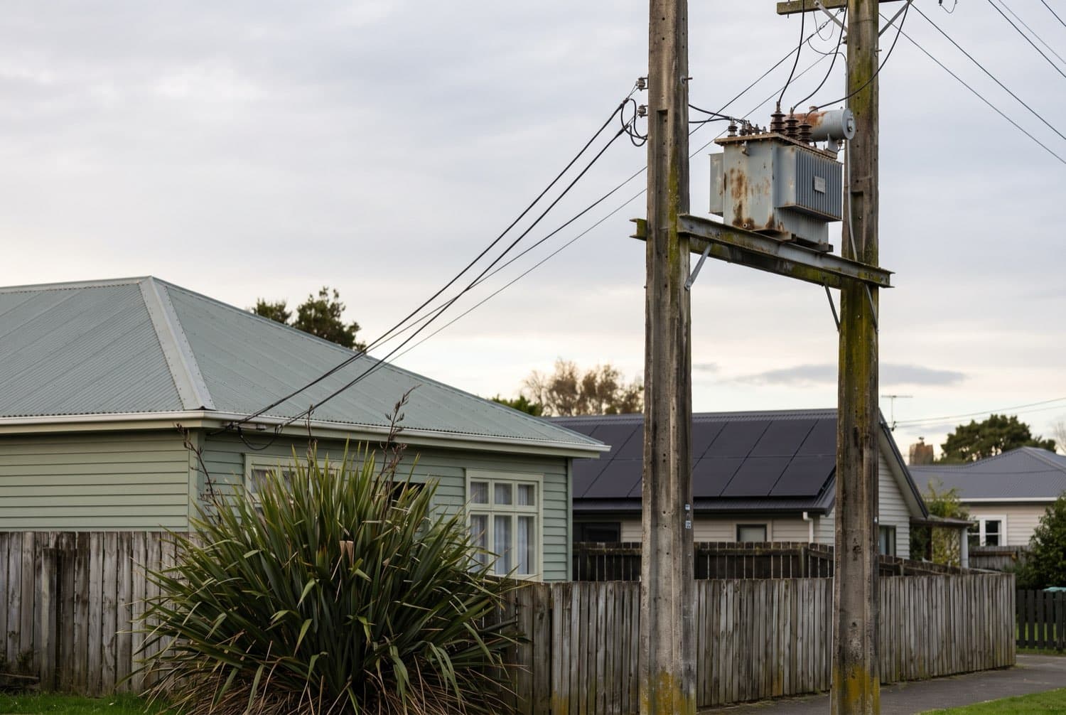A pole-top distribution transformer on a suburban New Zealand street with service wires running to a weatherboard house