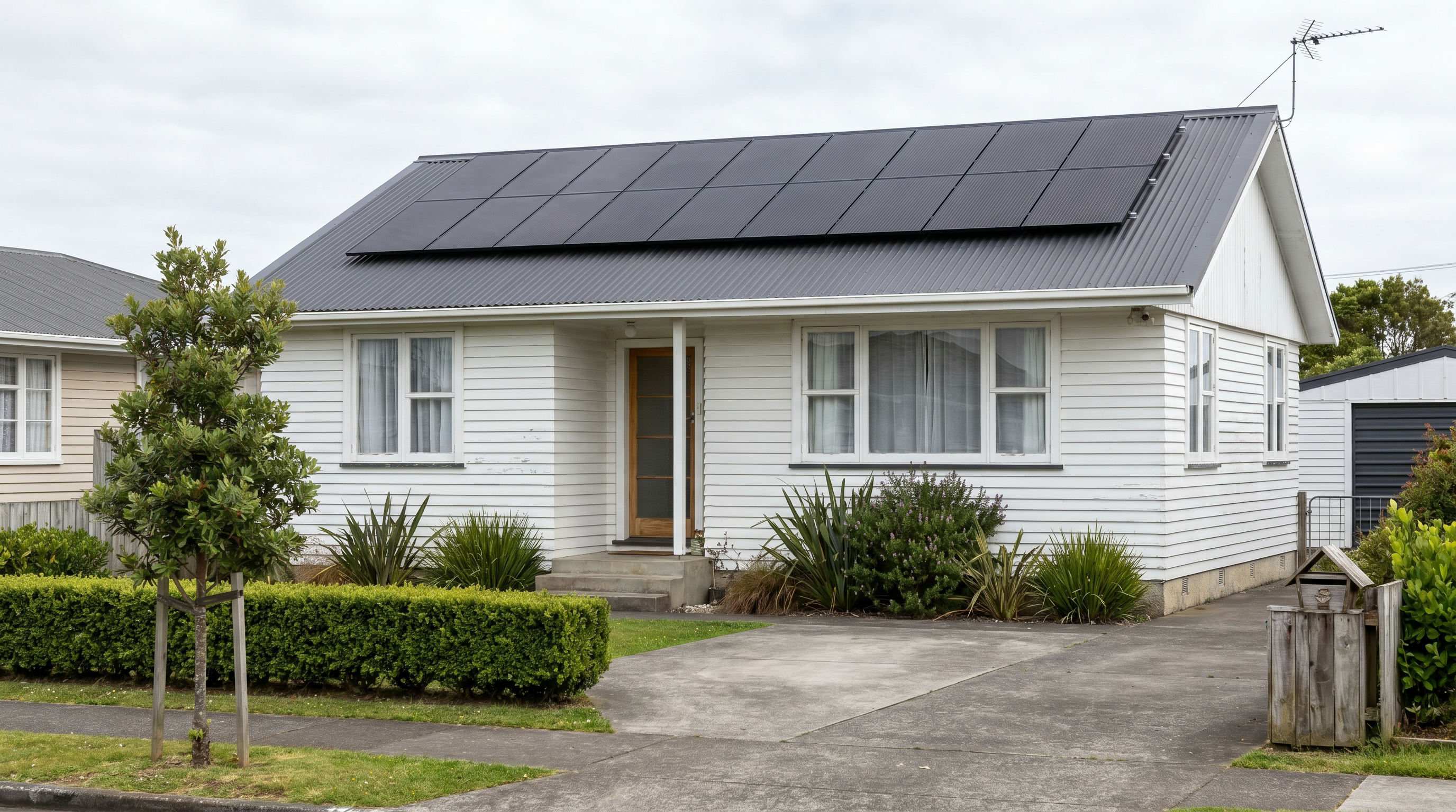 Documentary photo of a modest South Auckland white-weatherboard suburban home with a tidy row of all-black solar panels on its charcoal corrugated steel roof
